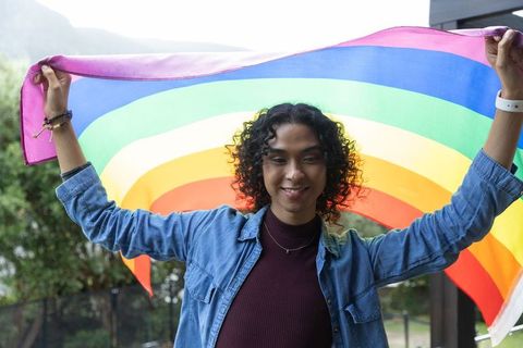 Non-binary adult holding rainbow flag across shoulders celebrating pride on balcony terrace