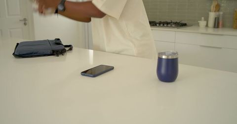 African american man reaching for smartphone on white kitchen counter with tumbler and bag
