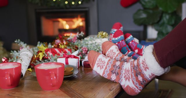 Two people wearing festive socks are by a cozy fireplace surrounded by holiday decorations and gifts. Two cups of hot chocolate are on the table, adding to the warm and relaxing atmosphere. This can be used for holiday-themed promotions, articles about Christmas traditions, or cozy wintertime settings.