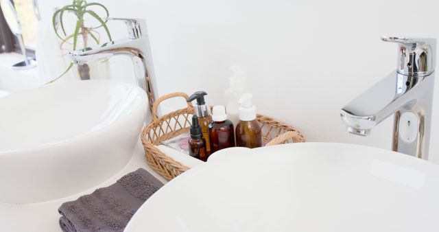 This image shows a clean and modern bathroom countertop featuring double white sinks with chrome faucets. A wicker basket containing various toiletries, such as bottles, is placed between the sinks. Gray towels are neatly folded next to the basket. This image can be used for home decor websites, lifestyle blogs, interior design inspiration, or advertisements for bathroom accessories.