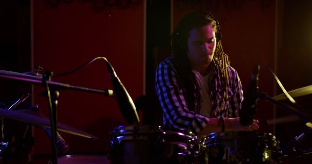 A biracial male artist playing drums, focusing on performance. He has long, dark hair, wearing striped shirt, in a dimly lit room