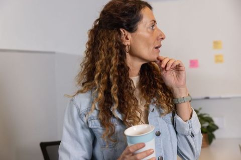 Woman standing in office holding coffee cup touching chin reviewing whiteboard notes