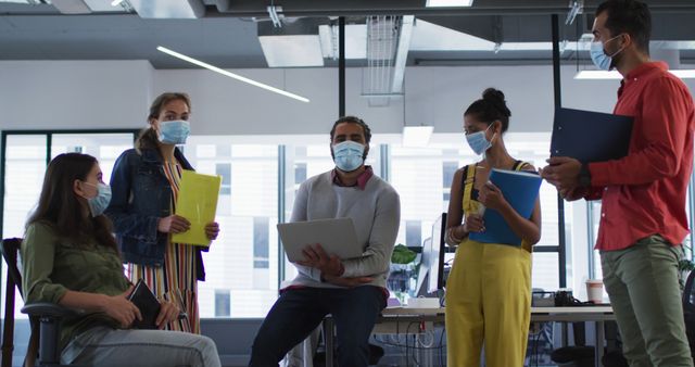 Portrait of diverse group of work colleagues wearing face mask holding laptop, tablet and documents. working at the office of an independent creative business during covid 19 coronavirus pandemic