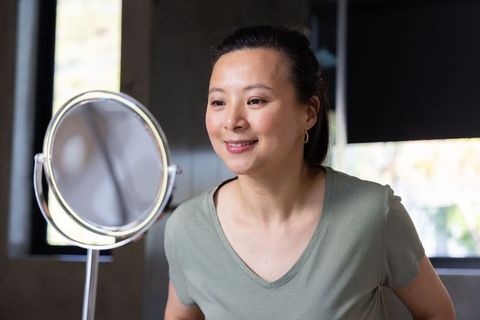 Mature asian woman smiling and inspecting reflection at vanity mirror during morning skincare routin
