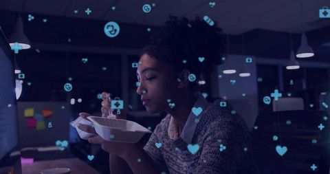 Young woman working late eating at desk watching monitor with floating health icons