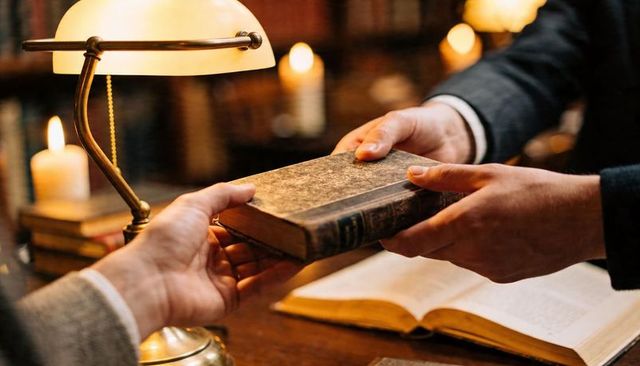 Passing antique hardcover book across wooden desk with brass lamp and open manuscript
