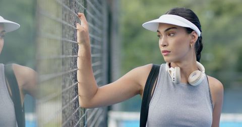 African american woman standing at tennis court fence wearing visor and headphones