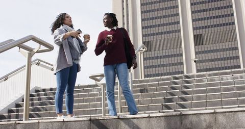 Multicultural young friends chatting on city steps holding coffee and smartphone