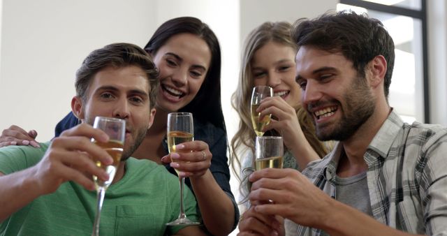 Group of friends toasting with glasses of champagne in a casual indoor setting. They are smiling, laughing, and enjoying each other's company. Perfect for themes of celebration, friendship, or social gatherings. This versatile image can be used in lifestyle blogs, social media posts, marketing materials, and advertisements related to beverages, events, or happy moments.