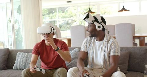 Diverse male friends laughing and playing VR on sofa with white headsets and controllers