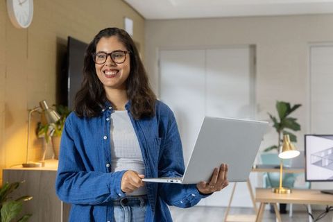 Indian woman holding laptop smiling in modern home office for remote work productivity