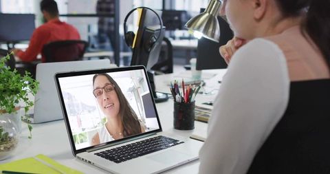 Female employee joining remote video call at modern open-plan office desk on laptop