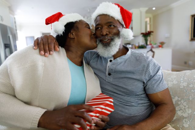 Front view of an old african-american couple sitting on the couch with the woman about to kiss her husband while holding her gift. both of them are wearing santa hats