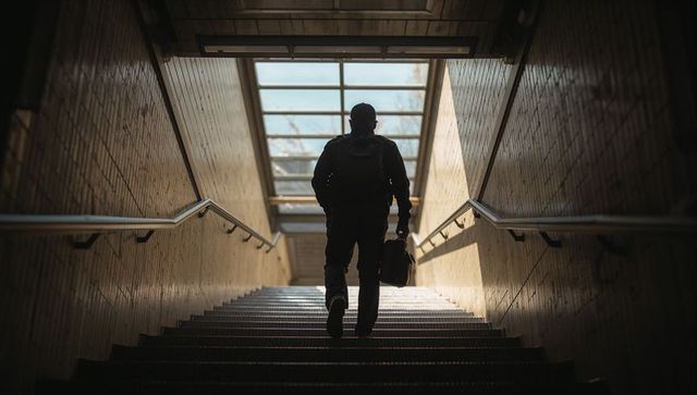 Silhouetted commuter climbing sunlit subway stairs carrying backpack and briefcase