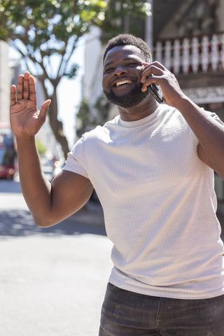 African man smiling and waving while talking on smartphone on sunlit city street