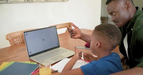 African american father tutoring son at wooden table pointing at laptop for homework help