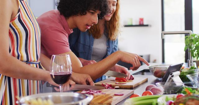 A group of friends is enthusiastically cooking together in a modern kitchen. Fresh vegetables are being chopped, with attention to detail on one friend's actions. A glass of red wine adds to the relaxed and enjoyable atmosphere. This picture can be used for content related to culinary arts, social gatherings, teamwork, cooking classes, and lifestyle blogs.