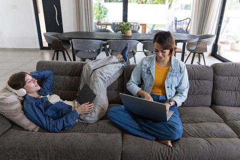 Friends relaxing on couch using laptop and tablet in modern open-plan living room, casual denim styl