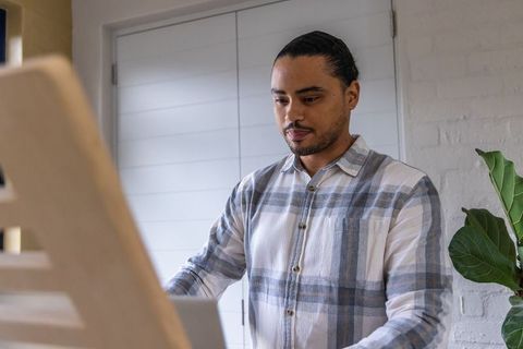Man Standing at Easel Desk Using Laptop in Minimalist Home Office with Plant