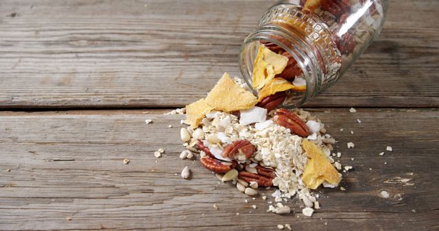 Spilled jar of granola on rustic wooden table top. Ideal for use in advertisements, websites, and articles promoting healthy eating, breakfast ideas, or homemade snacks. Perfect for visuals related to rustic or farmhouse kitchens.
