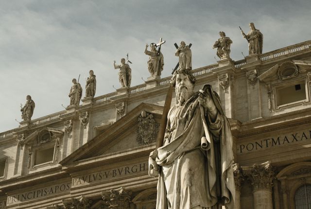 Prominent statue with detailed craftsmanship stands in front of St. Peter's Basilica. Other statues can be seen on top of the basilica, symbolizing religious significance and historical architecture. Ideal for use in travel blogs, history articles, educational pieces about Roman landmarks, or promotional material for tourism in Vatican City.