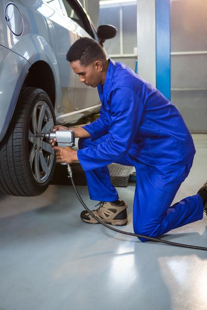 Mechanic wearing blue uniform kneeling and using a pneumatic wrench to fix a car wheel in a repair garage. Suitable for illustrating car maintenance, automotive repair services, and professional mechanics at work.