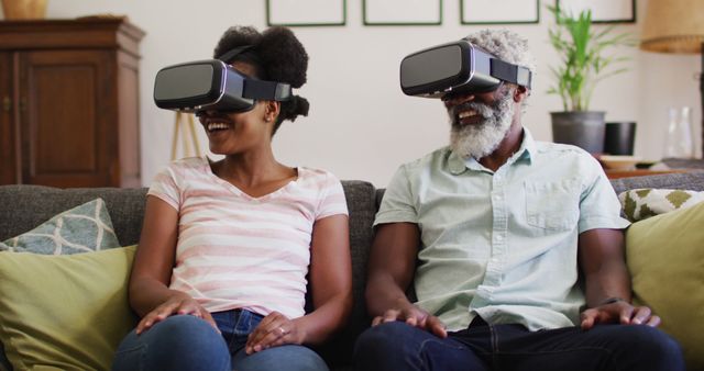 Father and daughter sitting on couch wearing VR headsets, smiling and enjoying the experience. This can be used for promoting virtual reality devices, showcasing family bonding with modern technology, or illustrating the impact of technology on home entertainment.