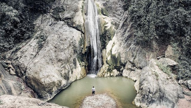 A solitary person is standing near a waterfall cascading down from rocky formations, surrounded by lush forest. Capturing a tranquil scene, this image depicts a serene connection with nature, ideal for themes of solitude, adventure, environmental consciousness, or travel. Suitable for use in travel blogs, nature magazines, stress relief articles, and promotional materials for outdoor activities or eco-tourism.