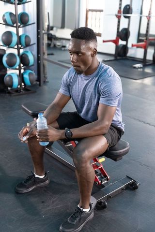 African american athlete sitting on bench holding water bottle resting after workout