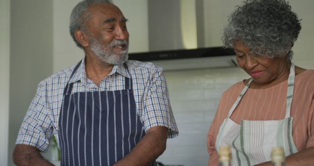 Senior couple enjoys cooking together in home kitchen, wearing aprons and smiling at each other, promoting ideas of enjoying shared activities in the domestic setting, perfect for retirement lifestyle content.
