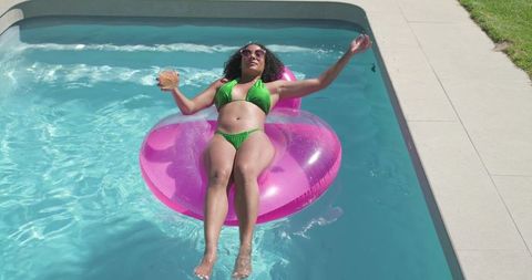 African american woman relaxing on pink inflatable ring, floating in pool with cocktail