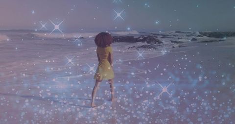 Woman standing on wet sand facing ocean in yellow dress with pastel bokeh and lens flares