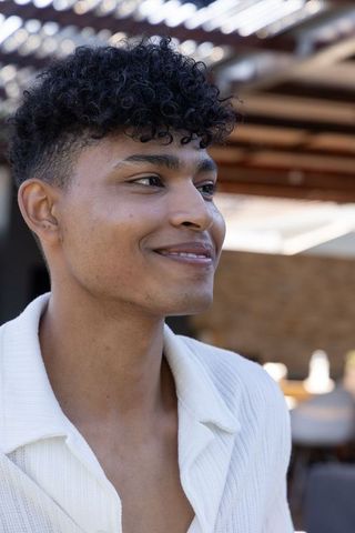 Sunlit african american man smiling under pergola on patio wearing white ribbed shirt