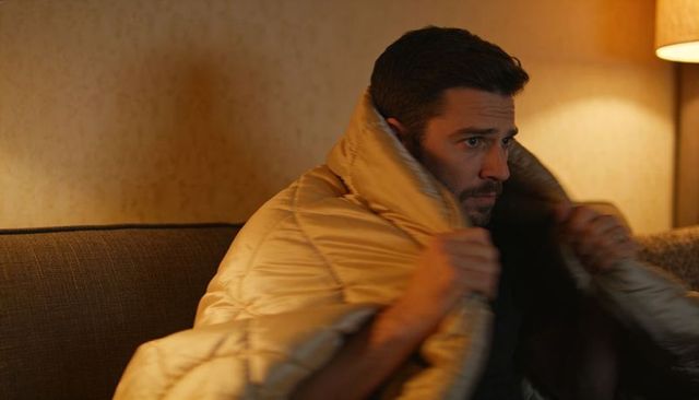 Man wrapping in quilted beige blanket on sofa at night, hugging for warmth and reflection