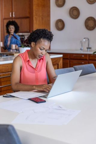 African american woman working on laptop at kitchen island with documents and smartphone