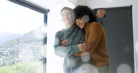 Embracing young couple by sunlit window wearing knit sweaters with hillside view