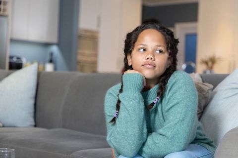 Thoughtful girl sitting on modern living room sofa wearing teal sweater and braided pigtails