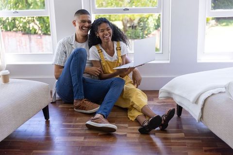 Young couple laughing while sitting on hardwood floor with laptop in bright modern home