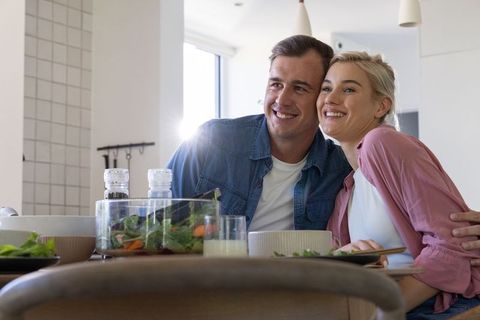 Couple smiling and sharing salad at sunlit modern kitchen table, cozy casual happy moment
