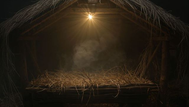 Empty rustic manger filled with straw under single hanging bulb in dark thatch stable