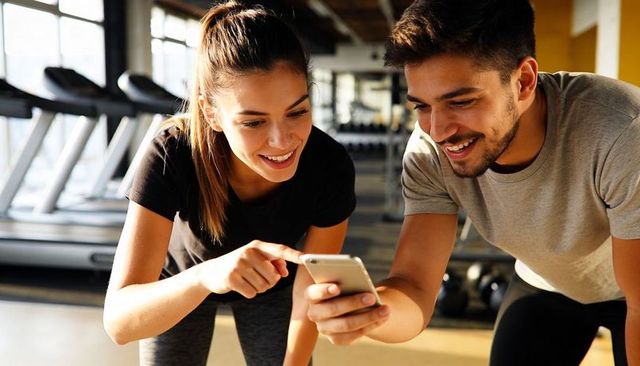 Smiling fitness partners checking smartphone during gym workout near treadmills and weights