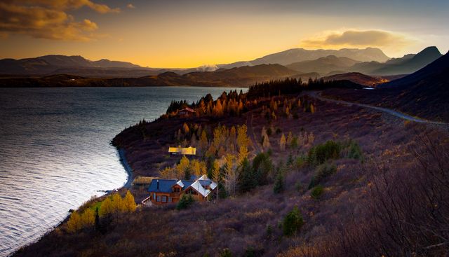This captivating mountain and lake landscape at sunset showcases a serene autumn scene with colorful foliage and cozy cabins. Ideal for travel brochures, nature-themed websites, inspirational posters, and desktop wallpapers emphasizing tranquility.