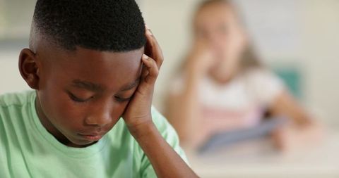 Pensive African boy leaning on hand at classroom desk while classmate holding tablet blurred