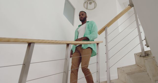 African American man stands confidently on stairs at home. His casual style and relaxed posture suggest comfort in his personal space.