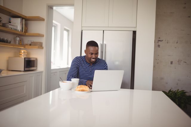Man sitting at kitchen counter using laptop while having breakfast. He is smiling and appears to be enjoying his morning routine. The kitchen is modern with white cabinets and stainless steel appliances. Ideal for use in articles about remote work, morning routines, technology in daily life, or modern home interiors.