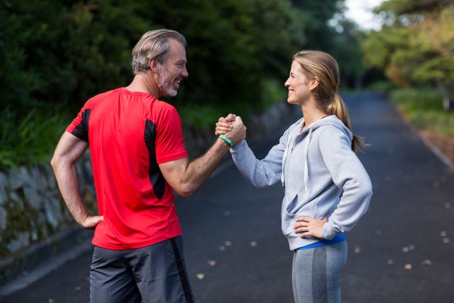 This image shows a smiling athletic couple holding hands on an open road, suggesting a strong bond and teamwork. They are dressed in sportswear, indicating they are engaged in outdoor exercise or a morning workout. This image can be used for promoting fitness, healthy living, and motivational content related to exercise and active lifestyles.