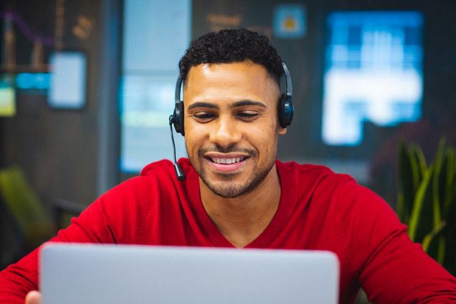 Young African American businessman wearing a headset, smiling while video conferencing in a modern office. Ideal for illustrating concepts of remote work, corporate communication, professional technology use, and modern business environments.