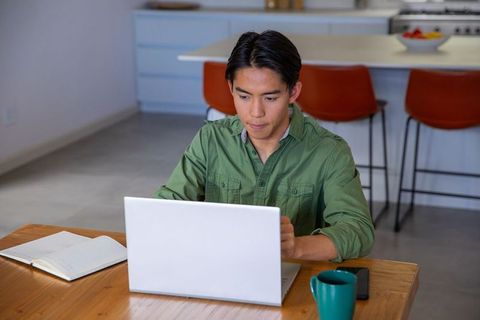 Young asian man working on laptop at modern kitchen table remote work study focused