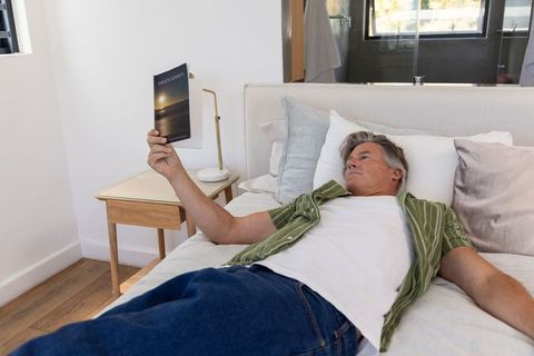 Middle-aged man relaxing on bed holding paperback book in bright modern bedroom wearing green shirt