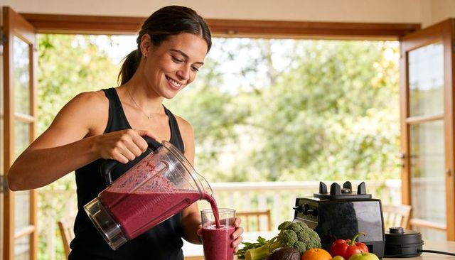 Smiling woman pouring berry smoothie from blender jar on sunlit kitchen terrace